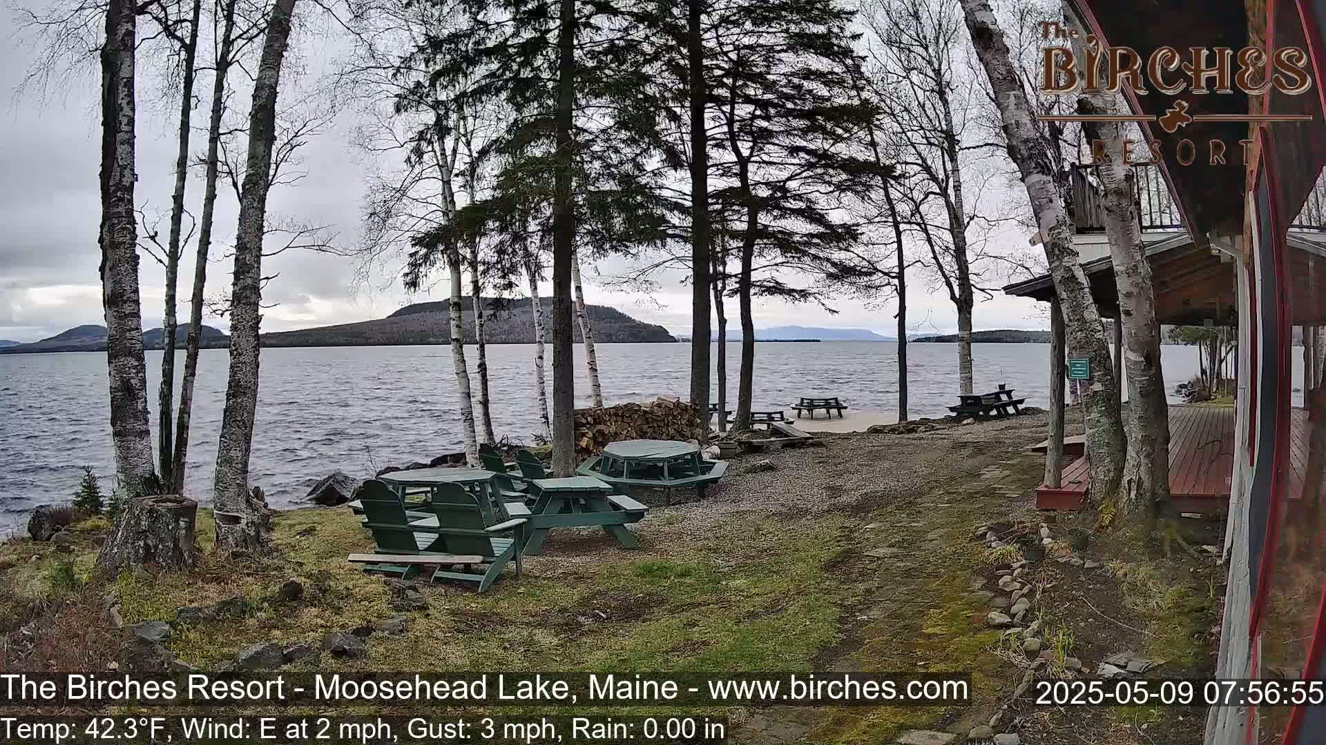 A lakeside view with several picnic tables and chairs under a cloudy sky, near a cabin, with bare trees and calm water.