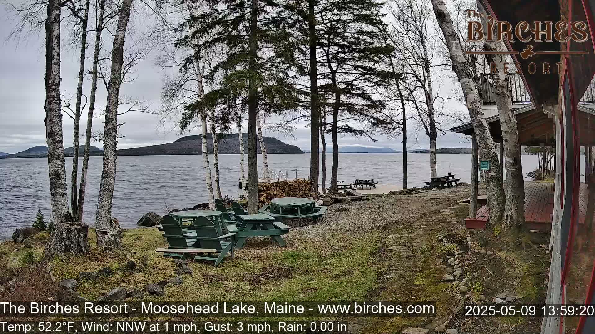 A lakeside resort with picnic tables and a wooden building, under a cloudy sky with light rain.