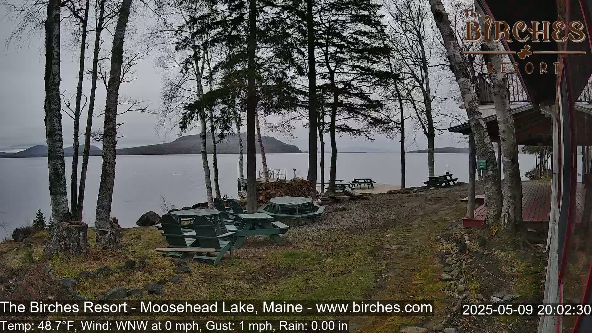 A lakeside resort area with several picnic tables and chairs, under a gray, overcast sky, overlooks a calm lake and distant hills.