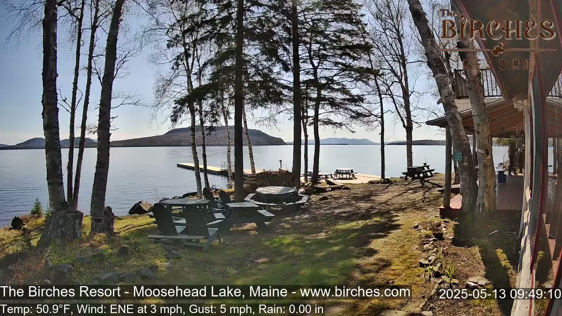 A sunny lakeside view shows picnic tables near a wooden building, a dock extending into calm water, and trees in the foreground and background, with hills visible across the lake.