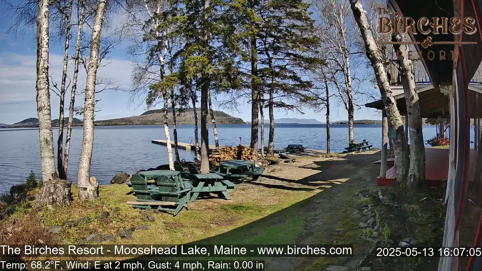 A sunny lakeside resort area with several picnic tables sits near a calm lake and a wooden building, surrounded by birch and evergreen trees.