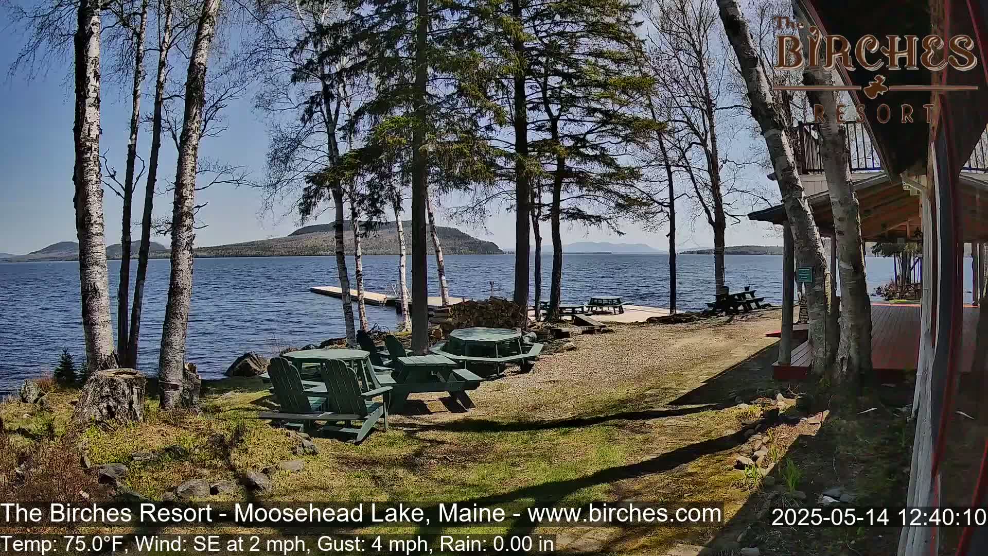 A lakeside patio with picnic tables and Adirondack chairs is situated near a building under a sunny sky, overlooking calm water and distant mountains.