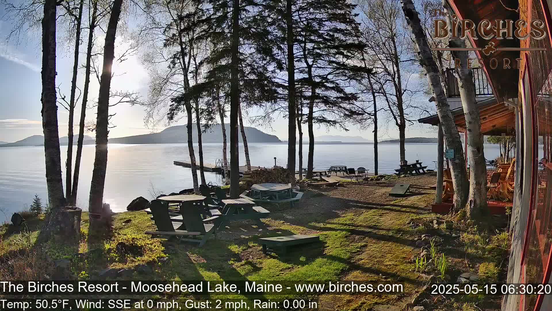 A sunny lakeside resort with picnic tables and a cornhole game, surrounded by birch trees, overlooks a calm lake and distant mountains.