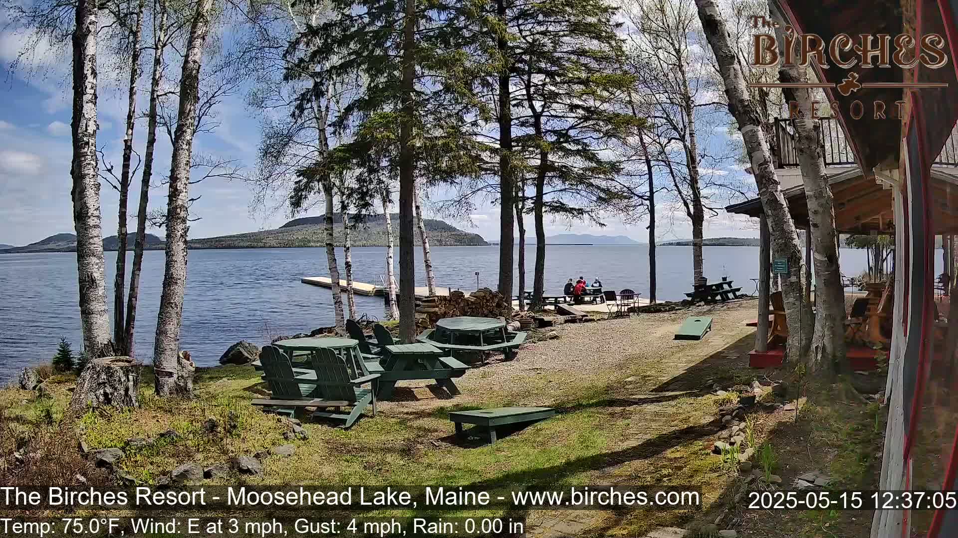 A lakeside resort area with picnic tables and Adirondack chairs, under a partly sunny sky, overlooks a calm lake and distant mountains.