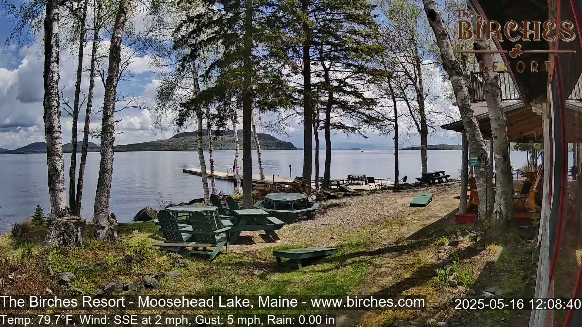 A lakeside resort with picnic tables and a cornhole game sits under a partly cloudy sky, with a calm lake and distant mountains visible in the background.