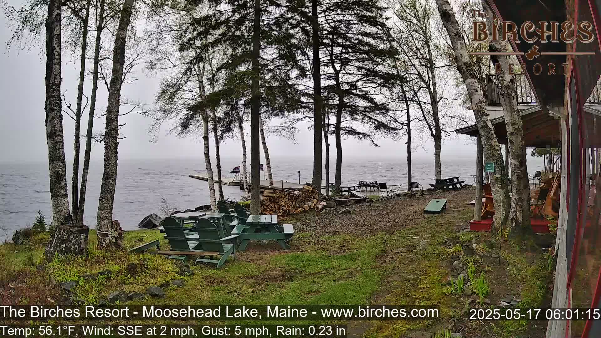 A lakeside patio with picnic tables and a cornhole game sits under a misty sky, near a wooden building and a dock extending into the water.