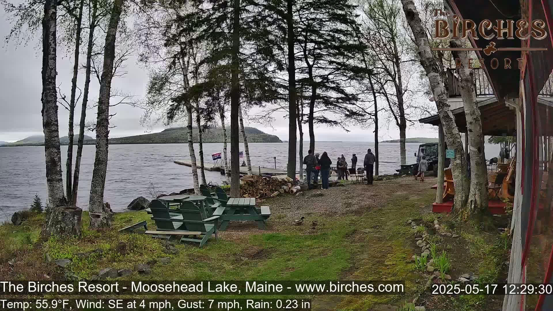 A lakeside resort scene shows several people gathered near a dock and picnic tables under a gray, rainy sky with a light wind.