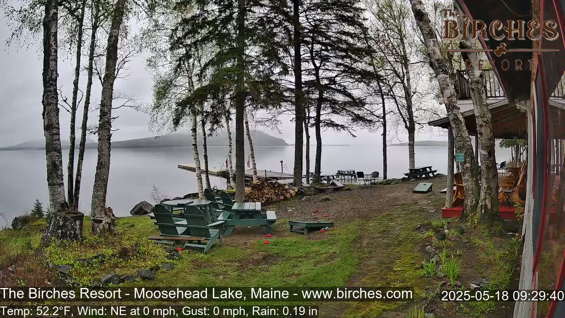 A lakeside resort with a gravel patio, picnic tables, Adirondack chairs, and a cornhole game, viewed on a misty, rainy day.