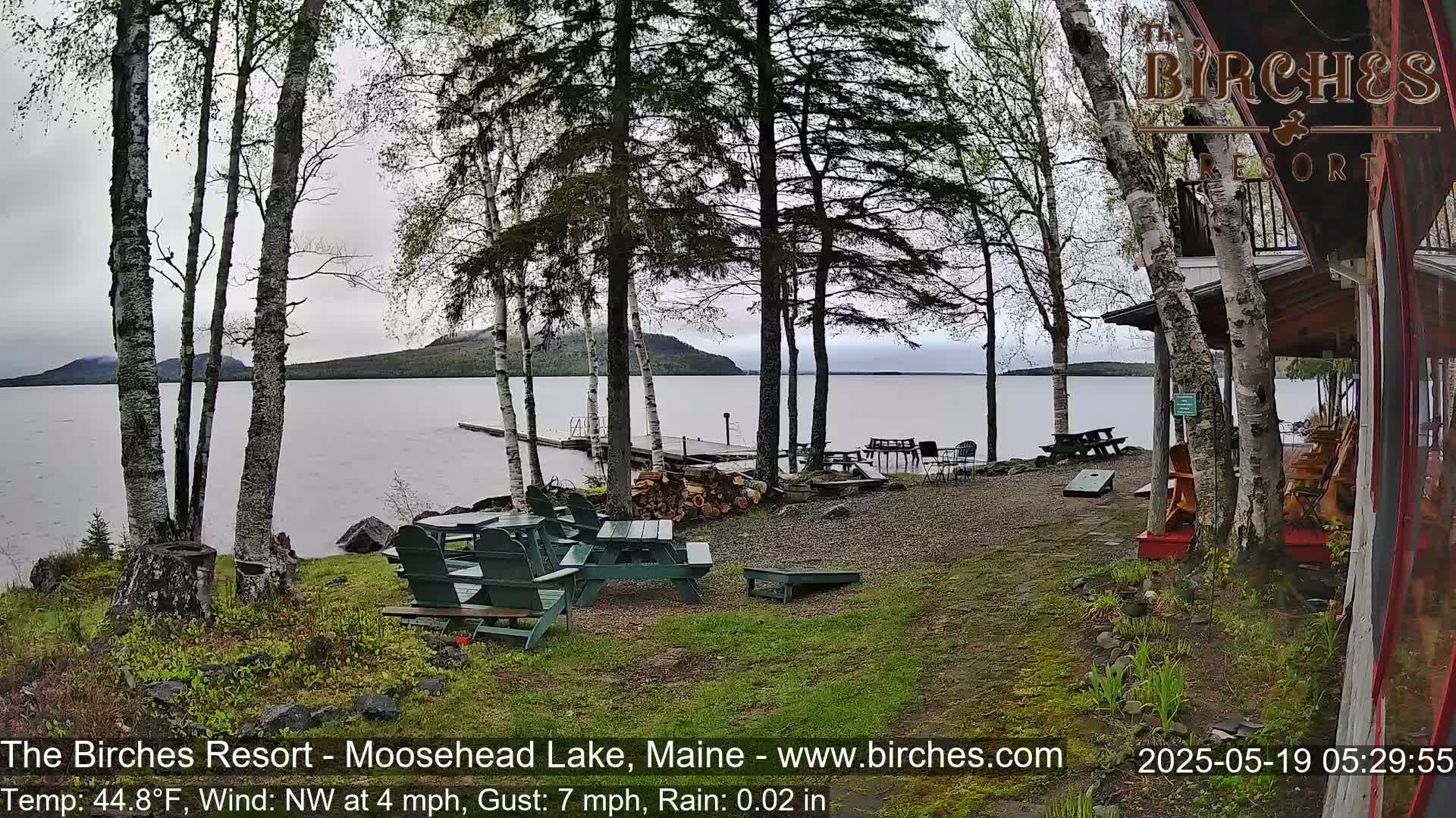 A lakeside patio with picnic tables and Adirondack chairs under a gray, rainy sky, overlooking a calm lake and distant hills.