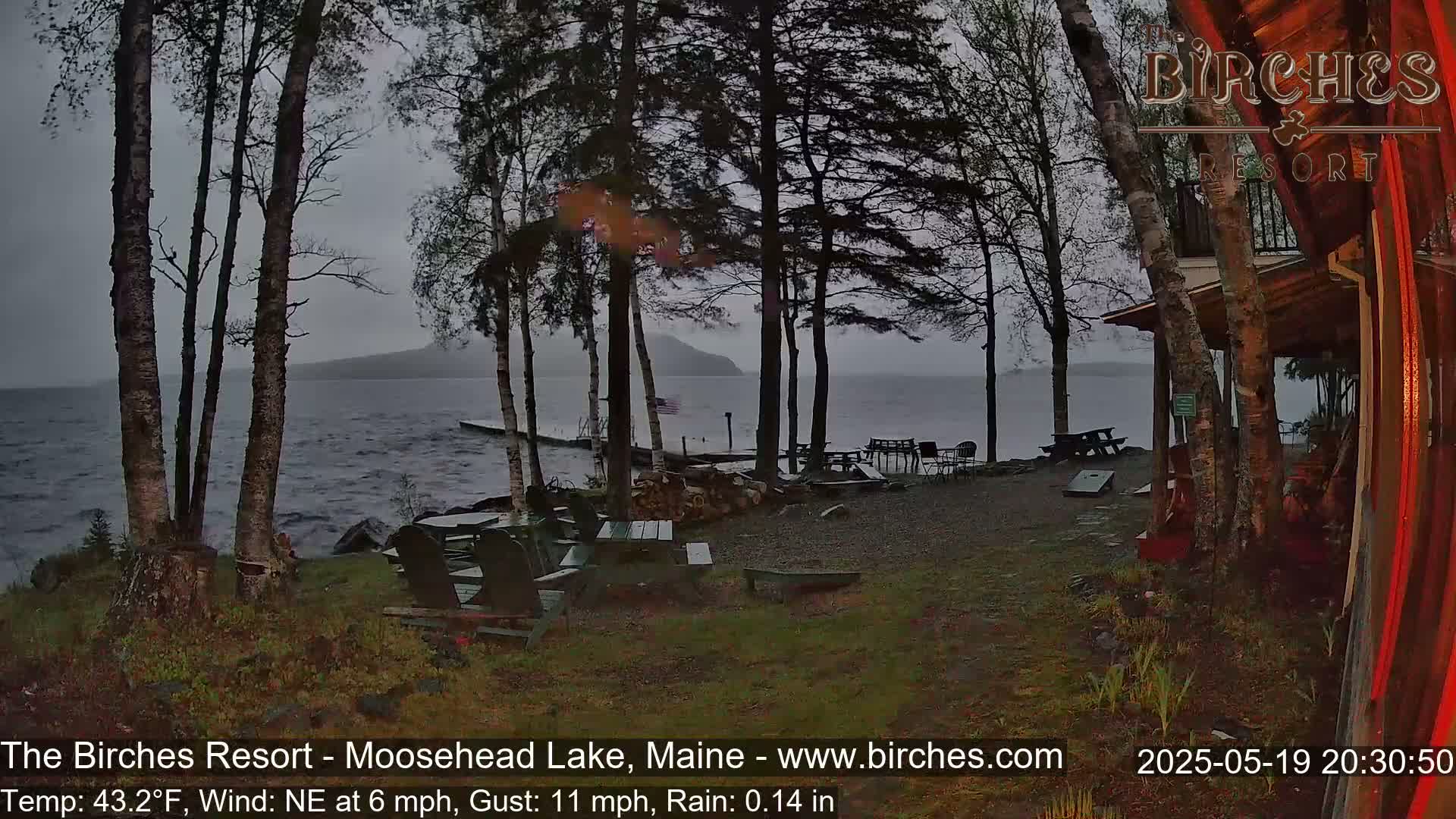 A lakeside resort with picnic tables and Adirondack chairs is seen under a rainy, overcast sky.