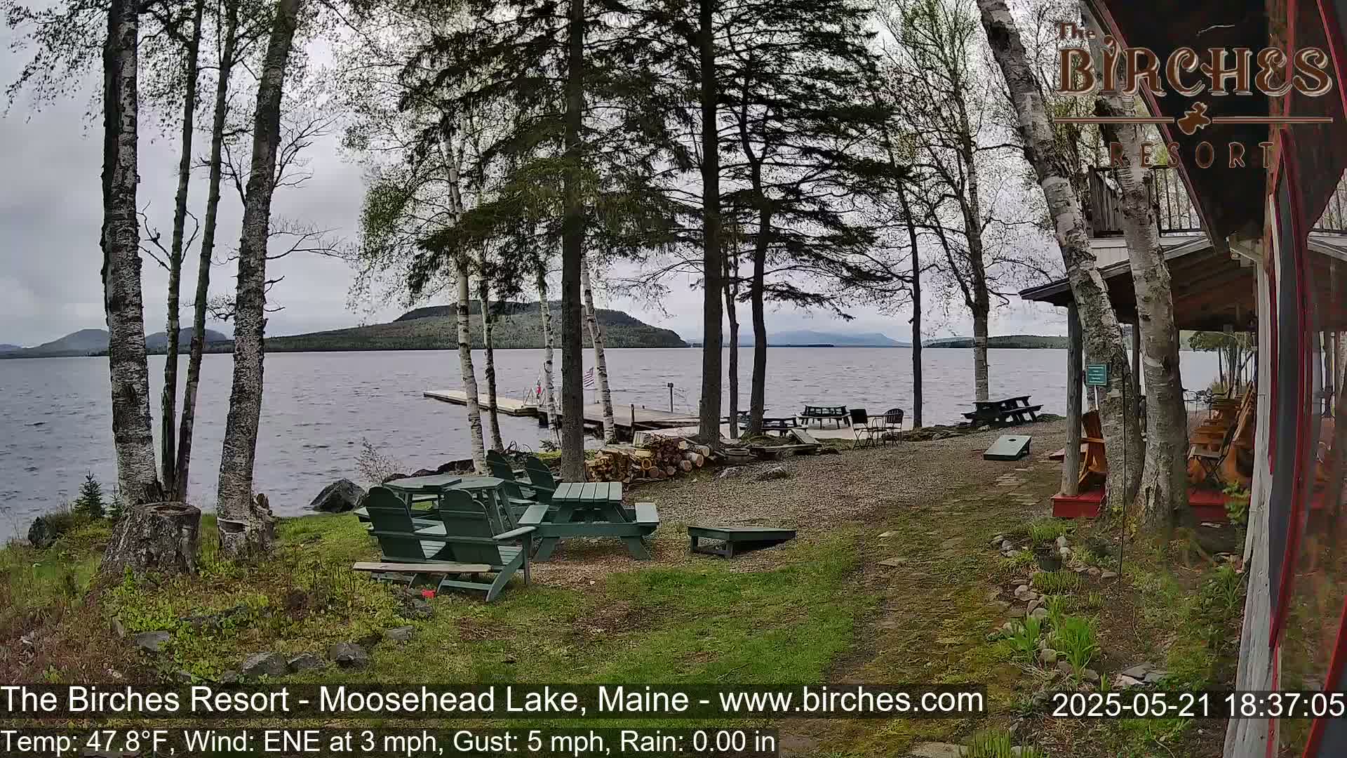 A lakeside resort area with picnic tables and Adirondack chairs under a cloudy sky, overlooking a calm lake with distant mountains.