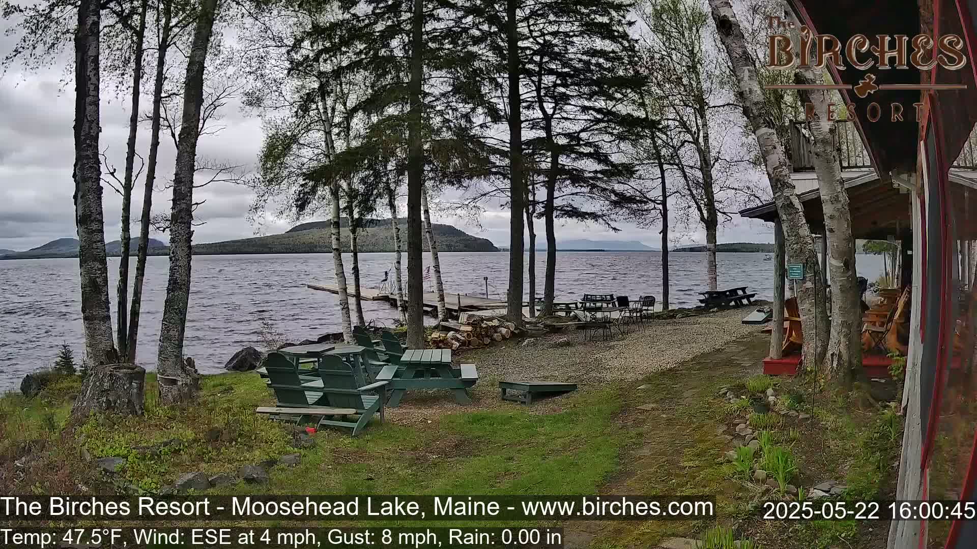 A lakeside patio with picnic tables and Adirondack chairs sits under a cloudy sky, overlooking a calm lake with a distant mountain range.