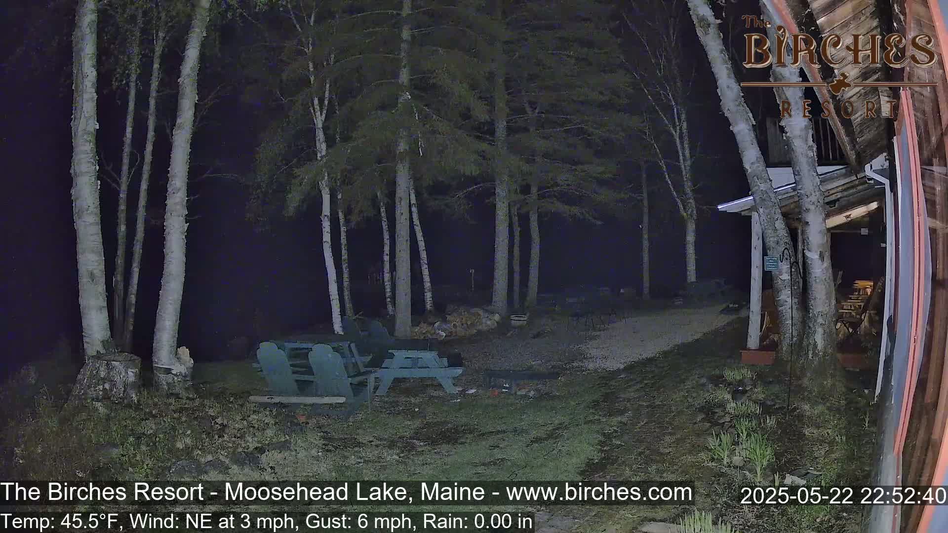 A nighttime outdoor scene shows a gravel area with picnic tables and a building partially obscured by birch trees under a dark sky with a northeast wind.