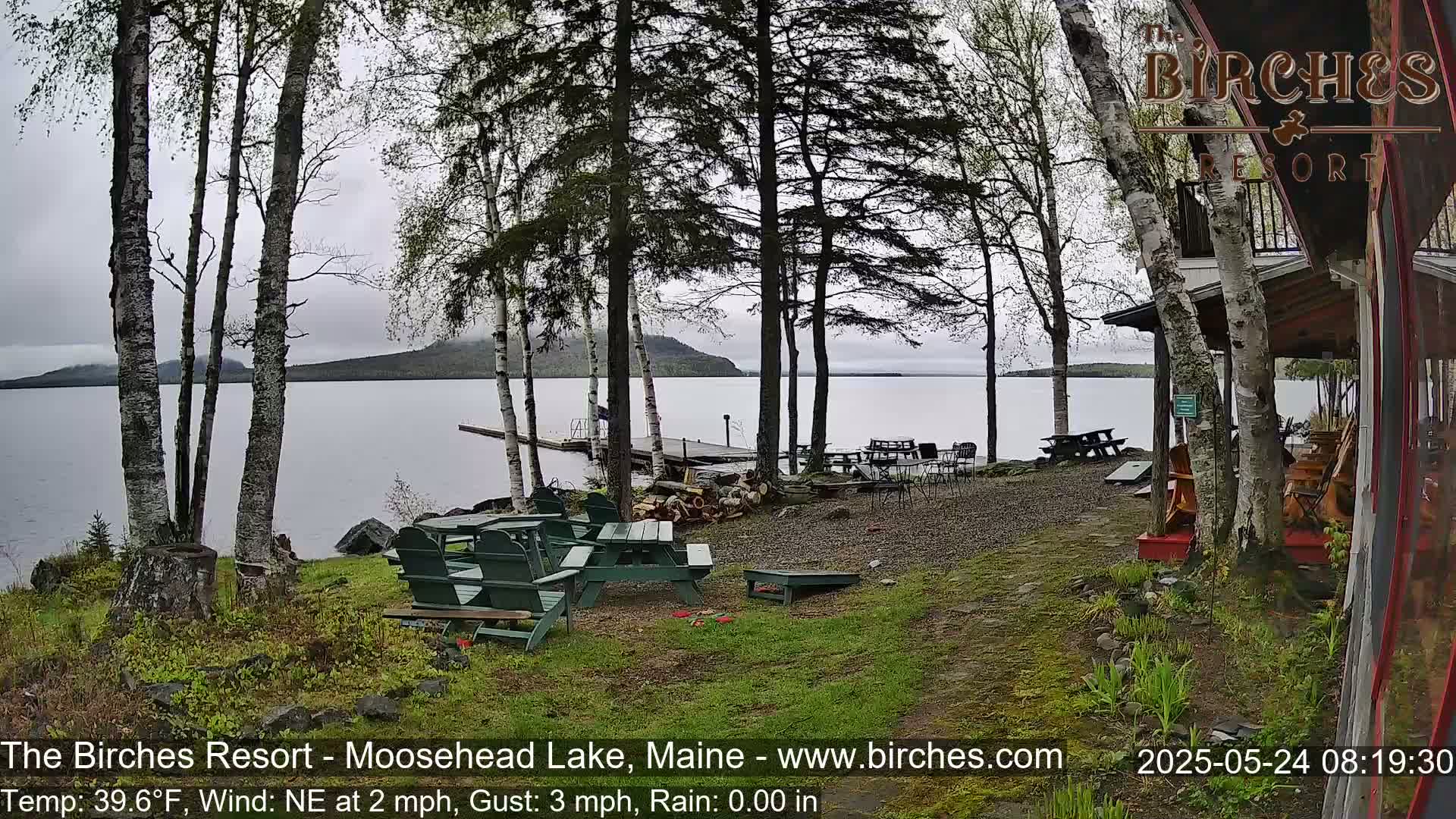 A lakeside resort area with several picnic tables and chairs sits under a mostly cloudy sky, with birch trees and a calm lake in the background.