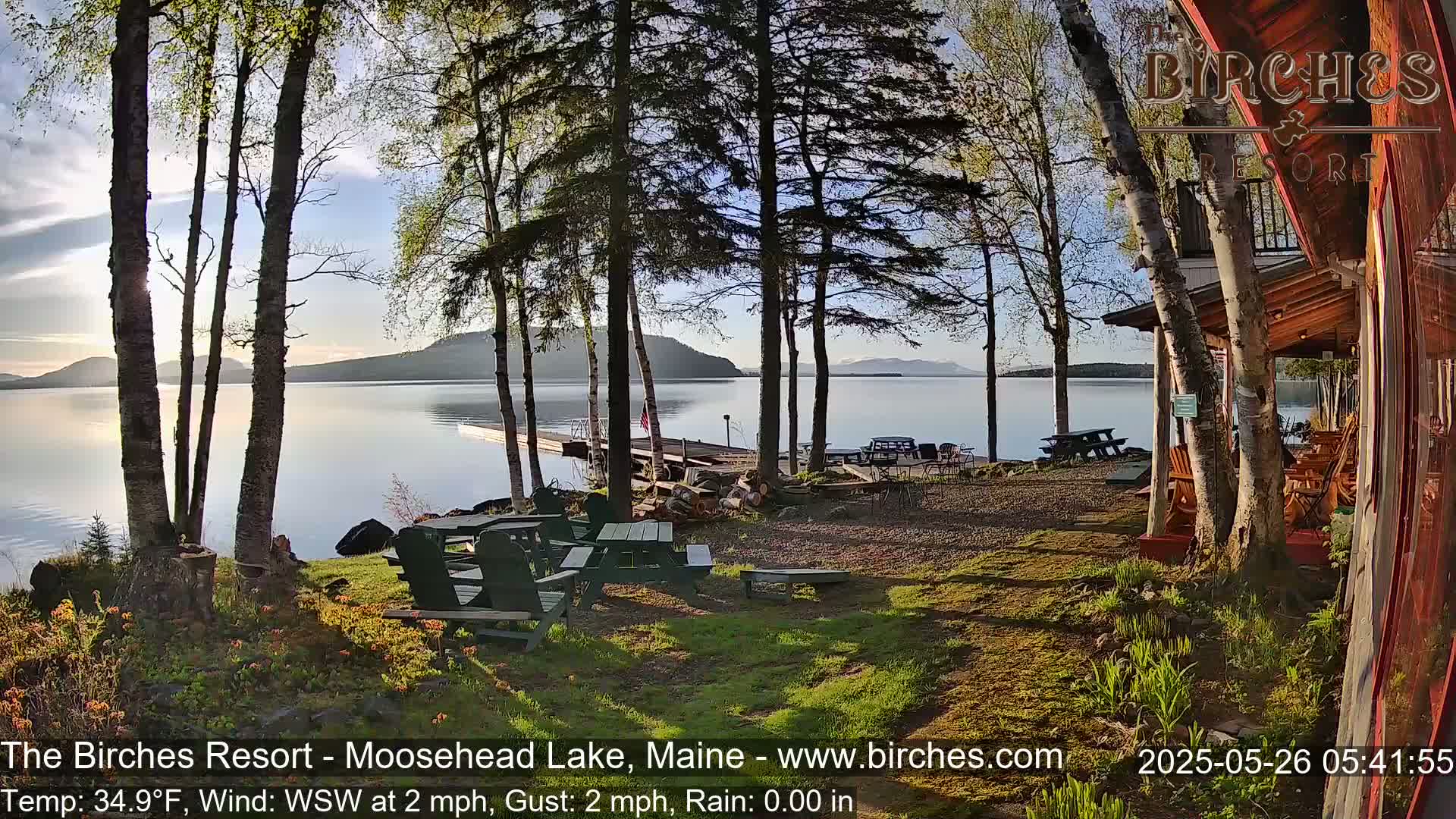 A lakeside patio with picnic tables and Adirondack chairs overlooks a calm lake and distant mountains under a partly cloudy, sunrise sky.