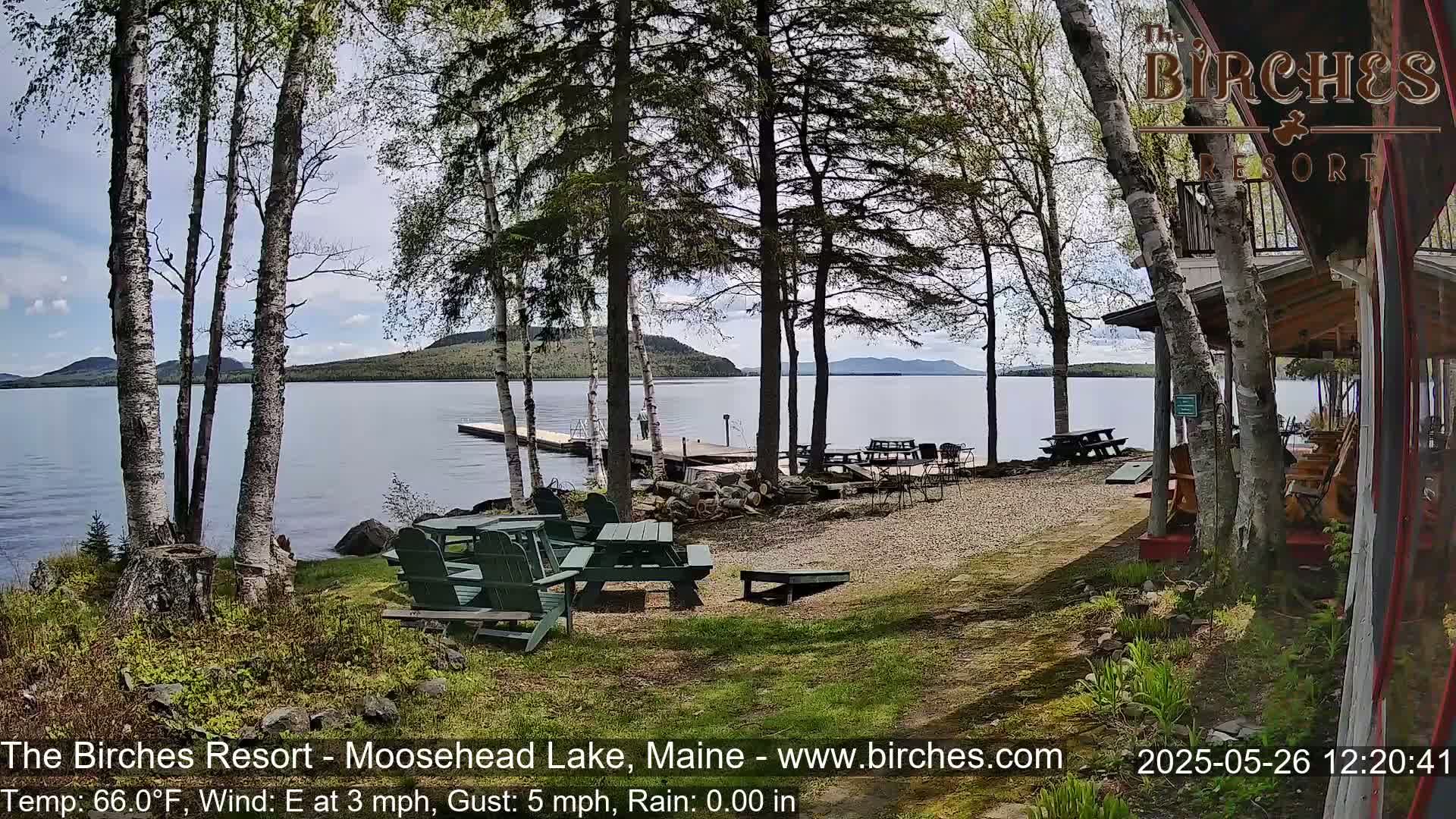 A lakeside resort area with picnic tables and Adirondack chairs overlooks a calm lake under a partly cloudy sky.