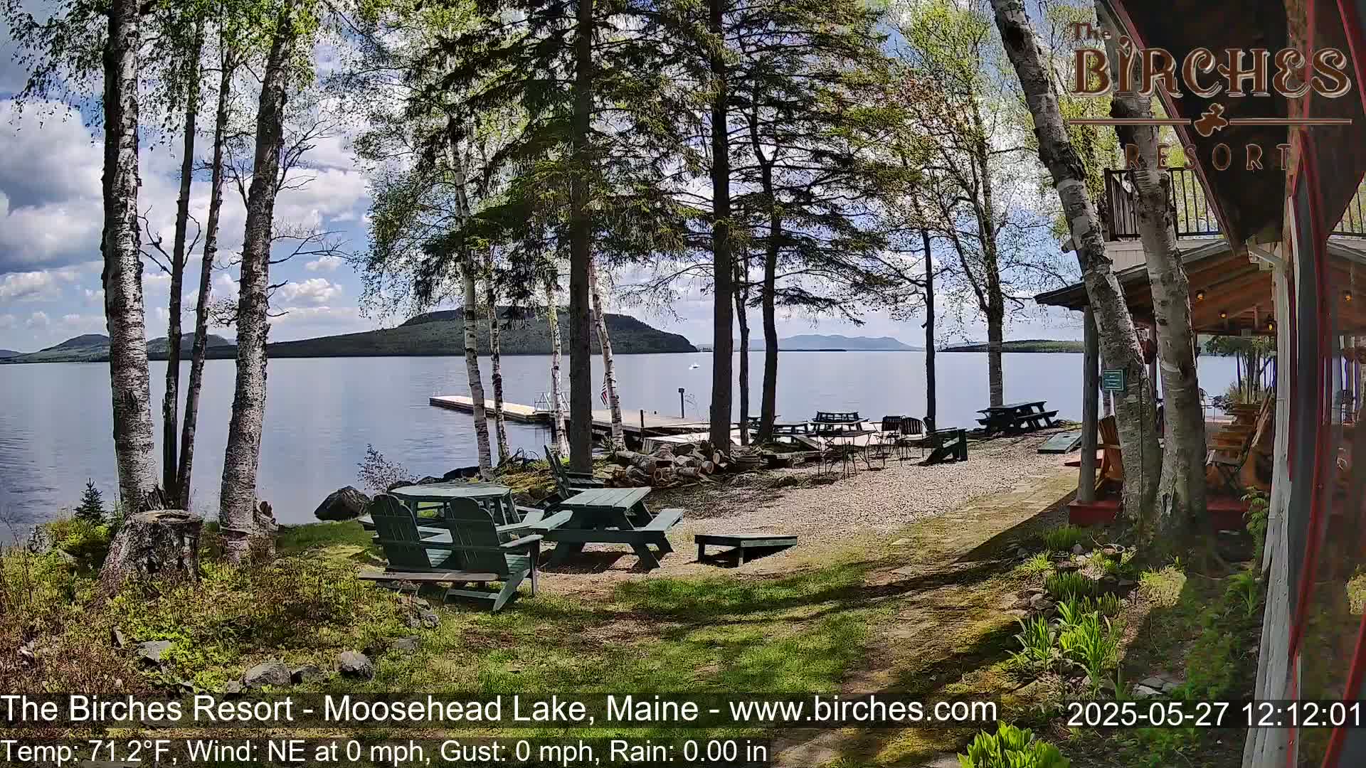 A sunny lakeside patio with picnic tables and Adirondack chairs overlooks a calm lake and distant hills under a partly cloudy sky.