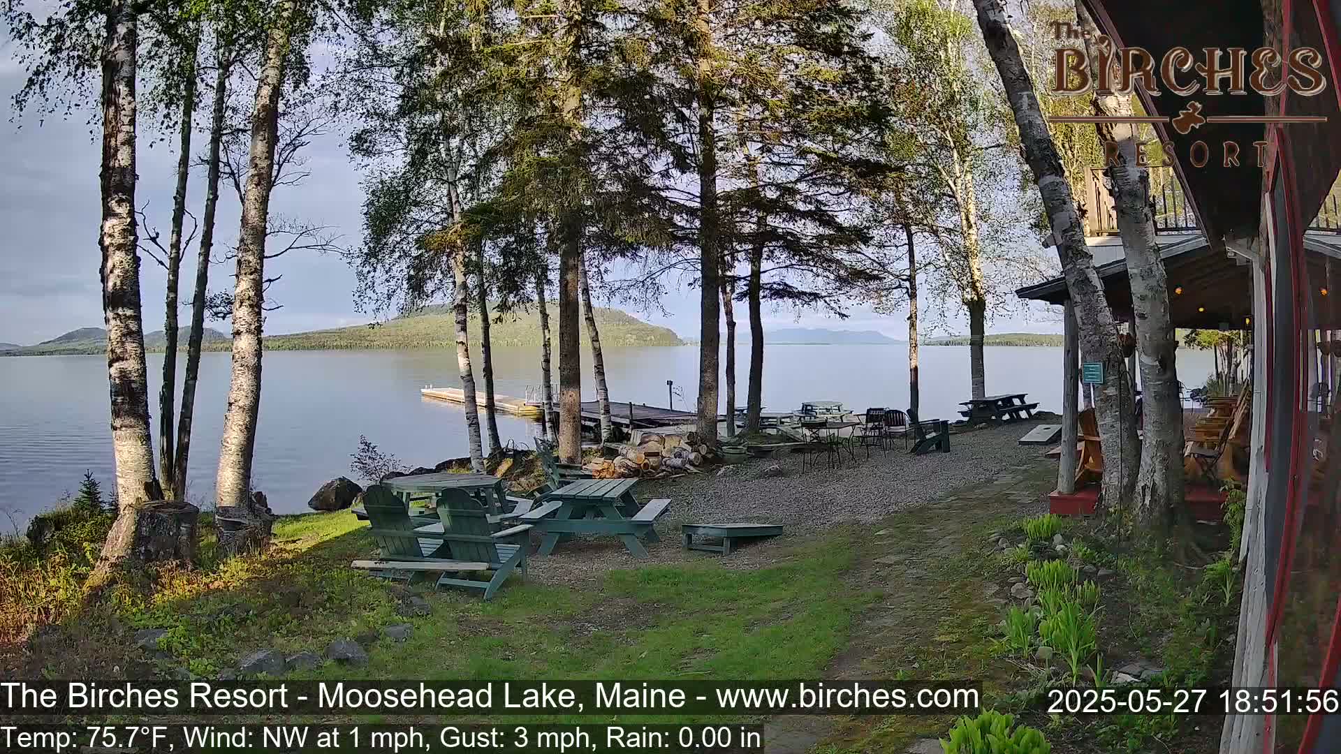 A lakeside resort with picnic tables and Adirondack chairs sits on a grassy area near a calm lake under a partly cloudy sky.