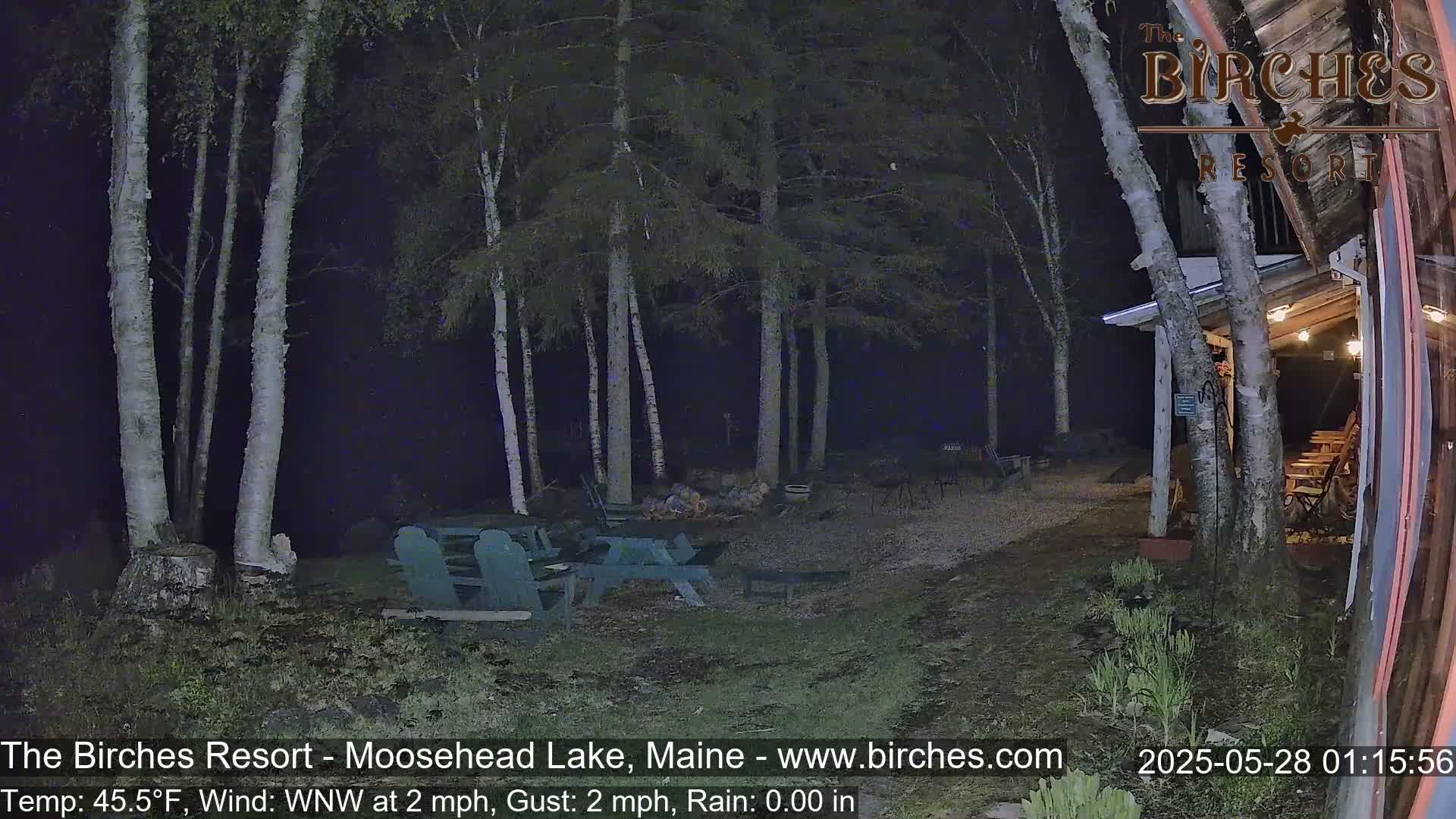 A nighttime outdoor scene shows a gravel area with picnic tables near birch trees and a building under a WNW wind at 2 mph with no rain.