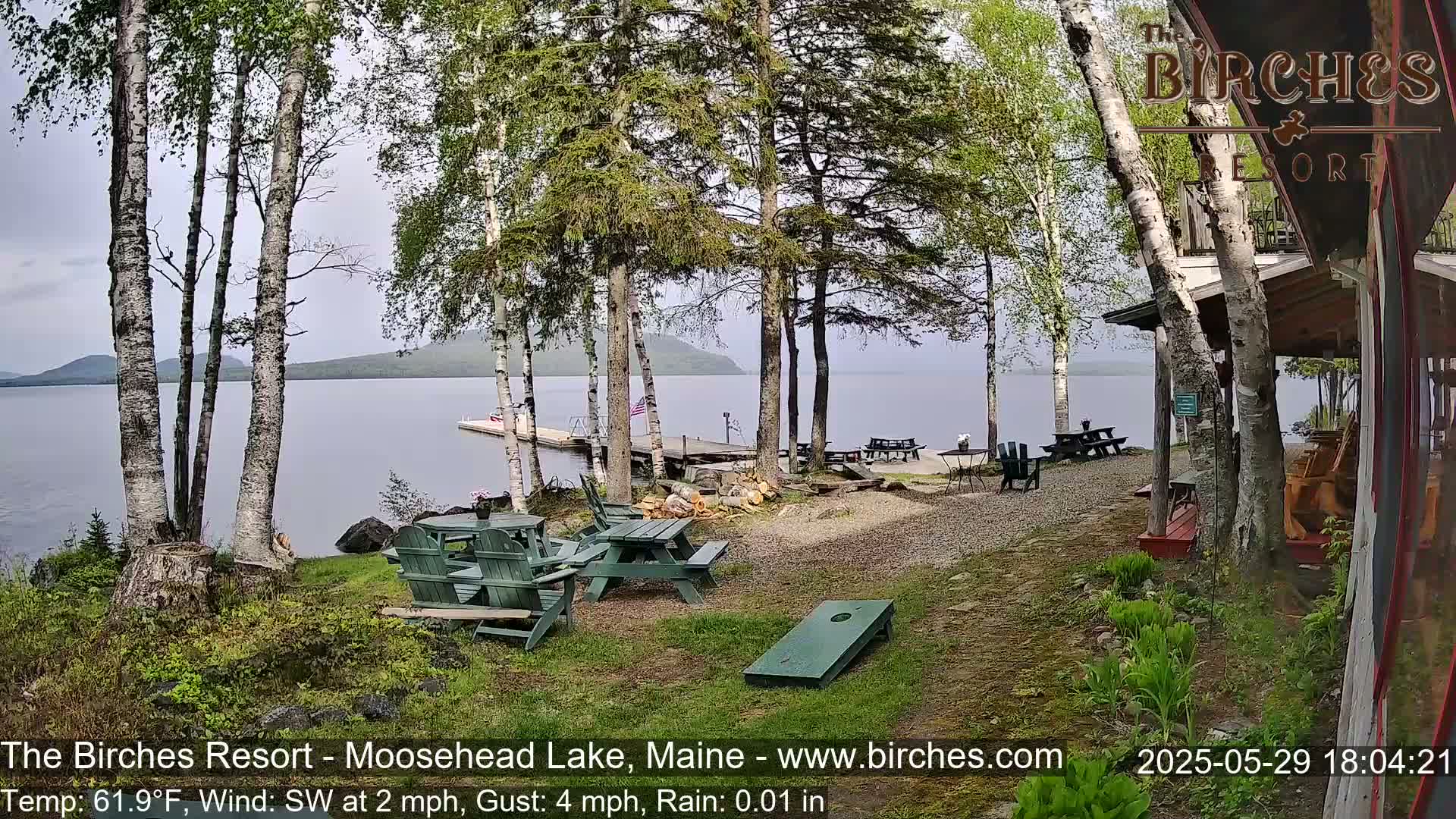 A lakeside patio area with picnic tables and Adirondack chairs, surrounded by birch trees, overlooks a calm lake under a mostly cloudy sky with light rain.