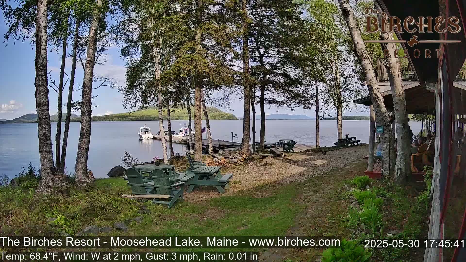 A lakeside resort scene with picnic tables, a dock with a boat, and birch trees under partly sunny skies with light rain.