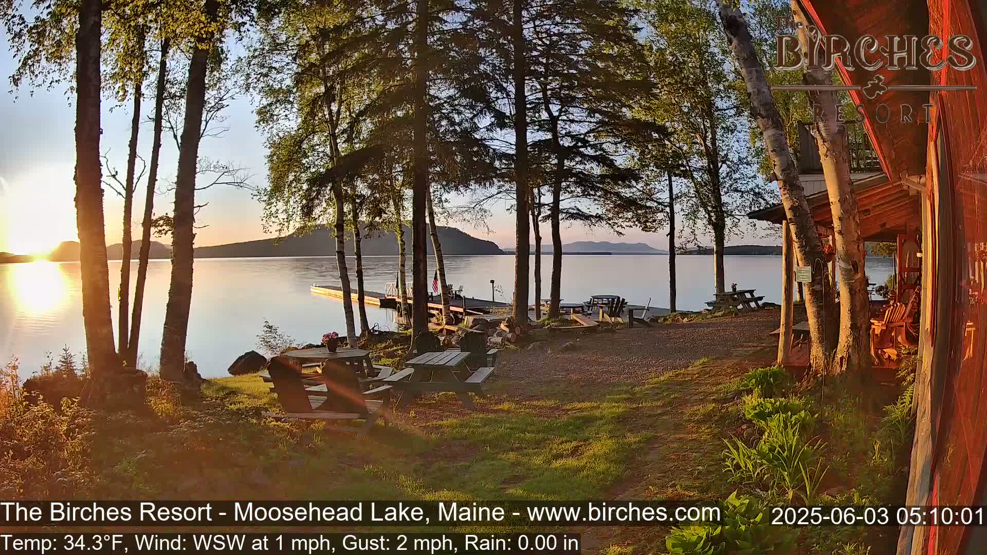 A lakeside resort at sunrise, with picnic tables and chairs situated near the water, under partly sunny skies and a light wind.