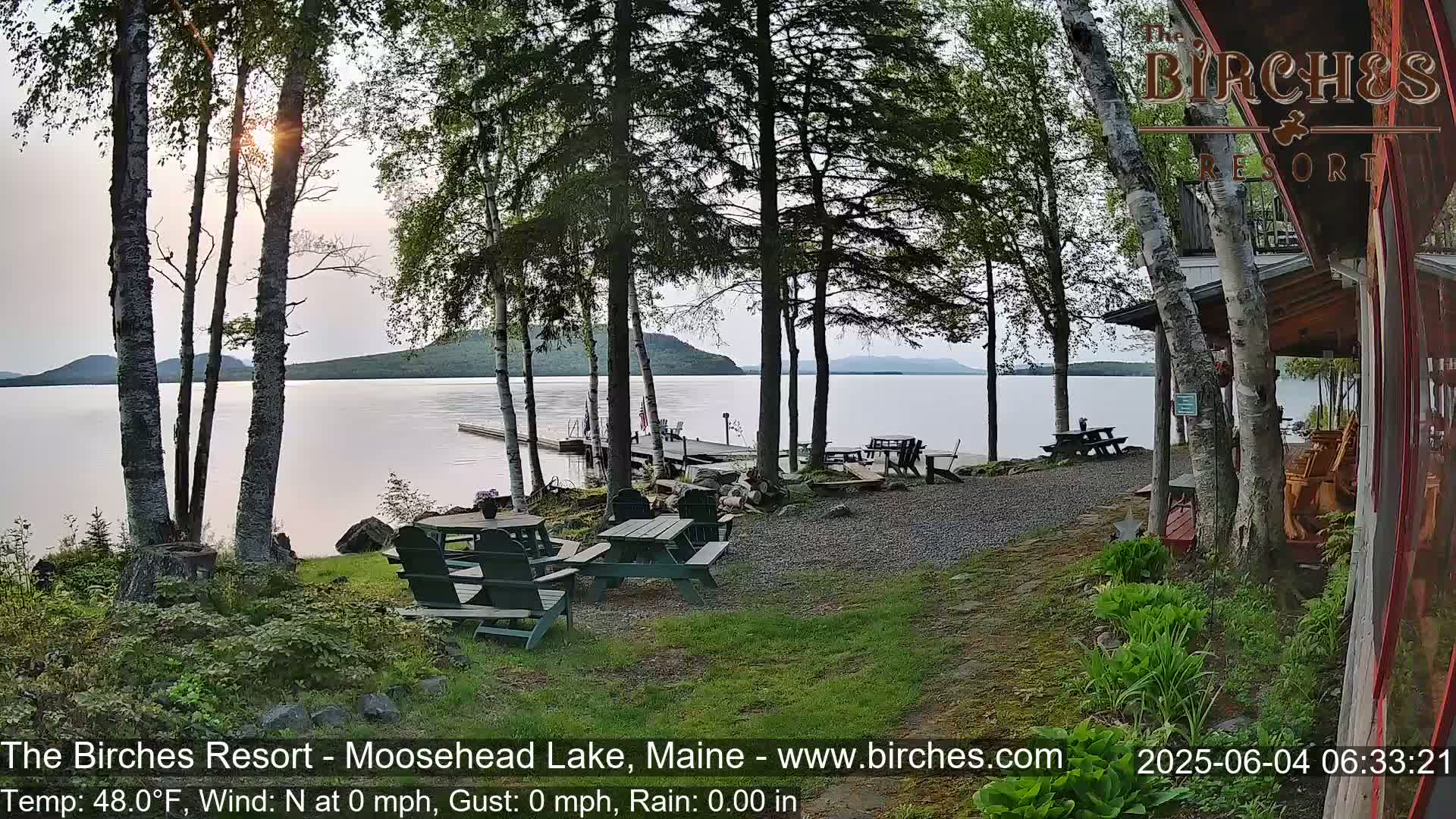 A calm lake view at sunset is seen from a lakeside resort with picnic tables and birch trees in the foreground.