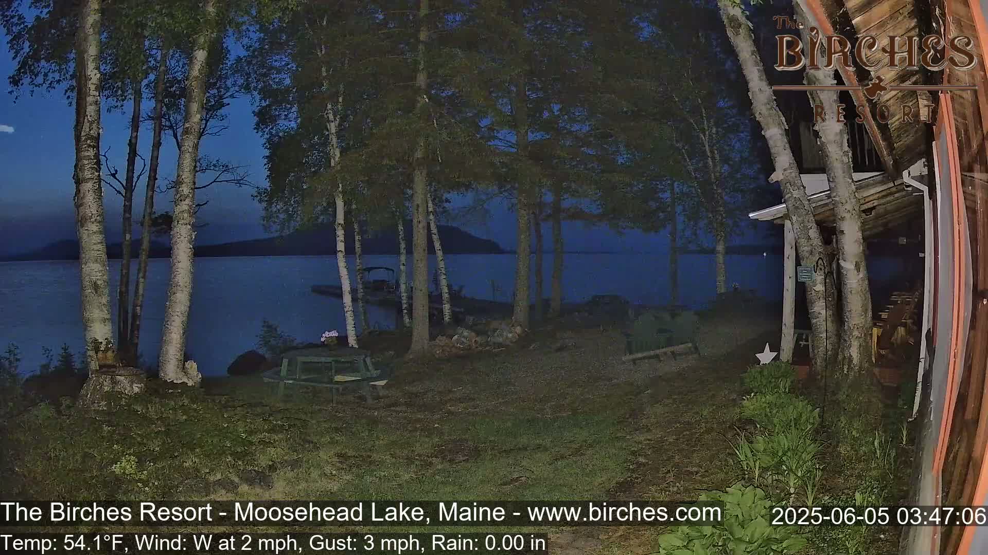 A dark, calm lake is seen at twilight through birch trees from a lakeside property, with a few picnic tables visible in the foreground.