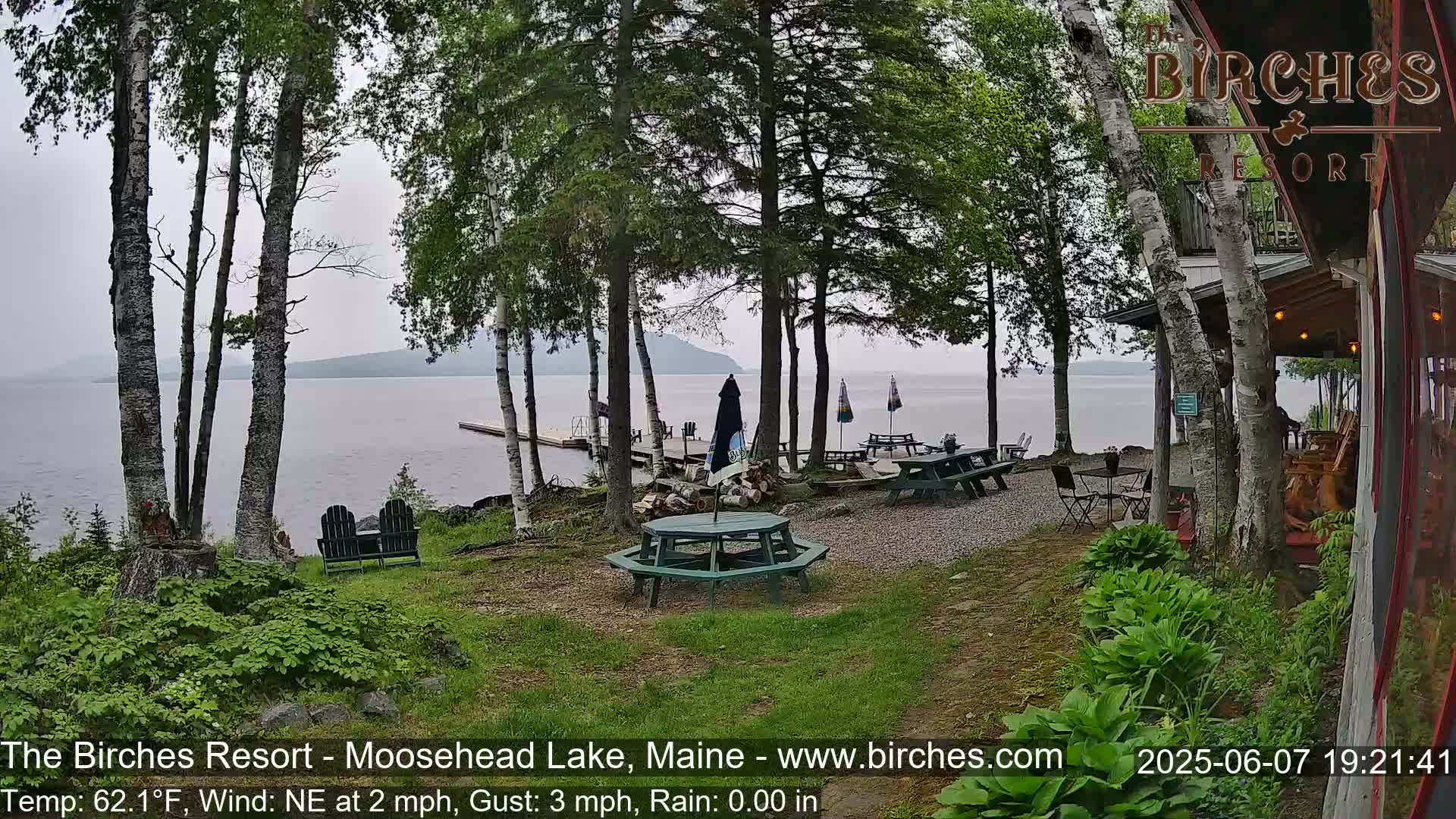 An outdoor lakeside patio area with picnic tables and chairs under a partly cloudy sky, near a calm lake with a dock and trees.