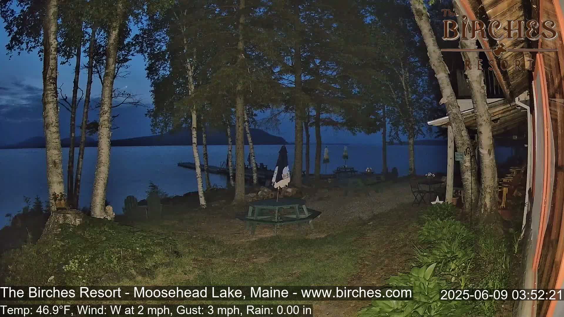 A lakeside picnic table sits under a twilight sky near birch trees and a building, with calm water visible in the background.