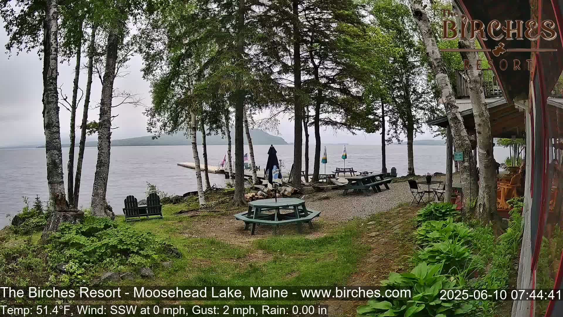An outdoor lakeside patio area with picnic tables and chairs sits under a cloudy sky beside a calm lake and several birch trees.