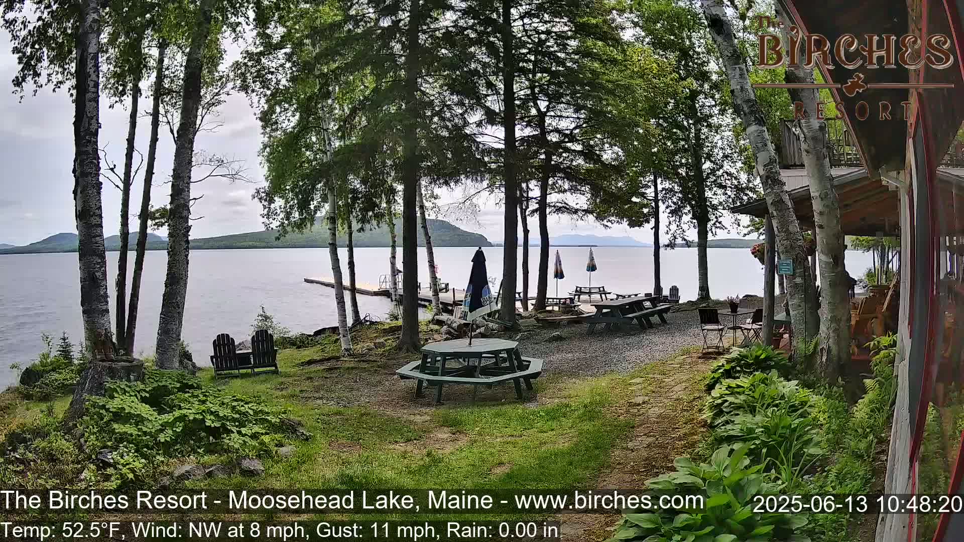 A lakeside resort with picnic tables and chairs under a cloudy sky, with a calm lake and distant mountains in the background.