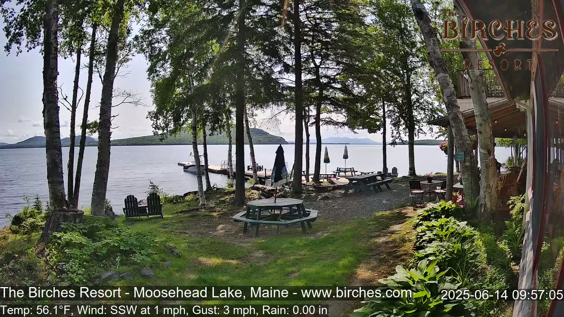 A lakeside patio with picnic tables and chairs is nestled among trees, overlooking a calm lake and distant mountains under partly cloudy skies.