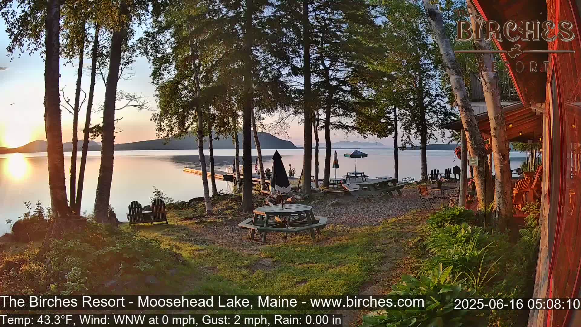 A calm lakeside scene at sunset shows picnic tables and chairs near a dock, surrounded by trees and a building, with the sun reflecting on the water.