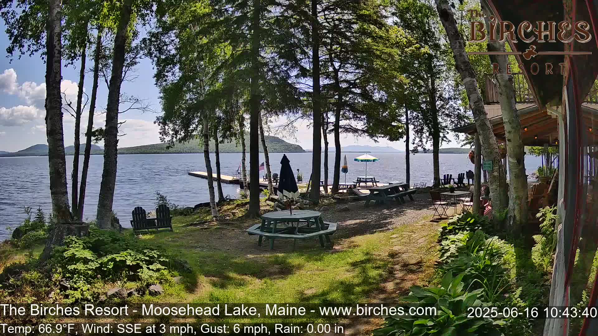 A lakeside resort area with picnic tables, Adirondack chairs, and a dock on a sunny day with a slight wind.