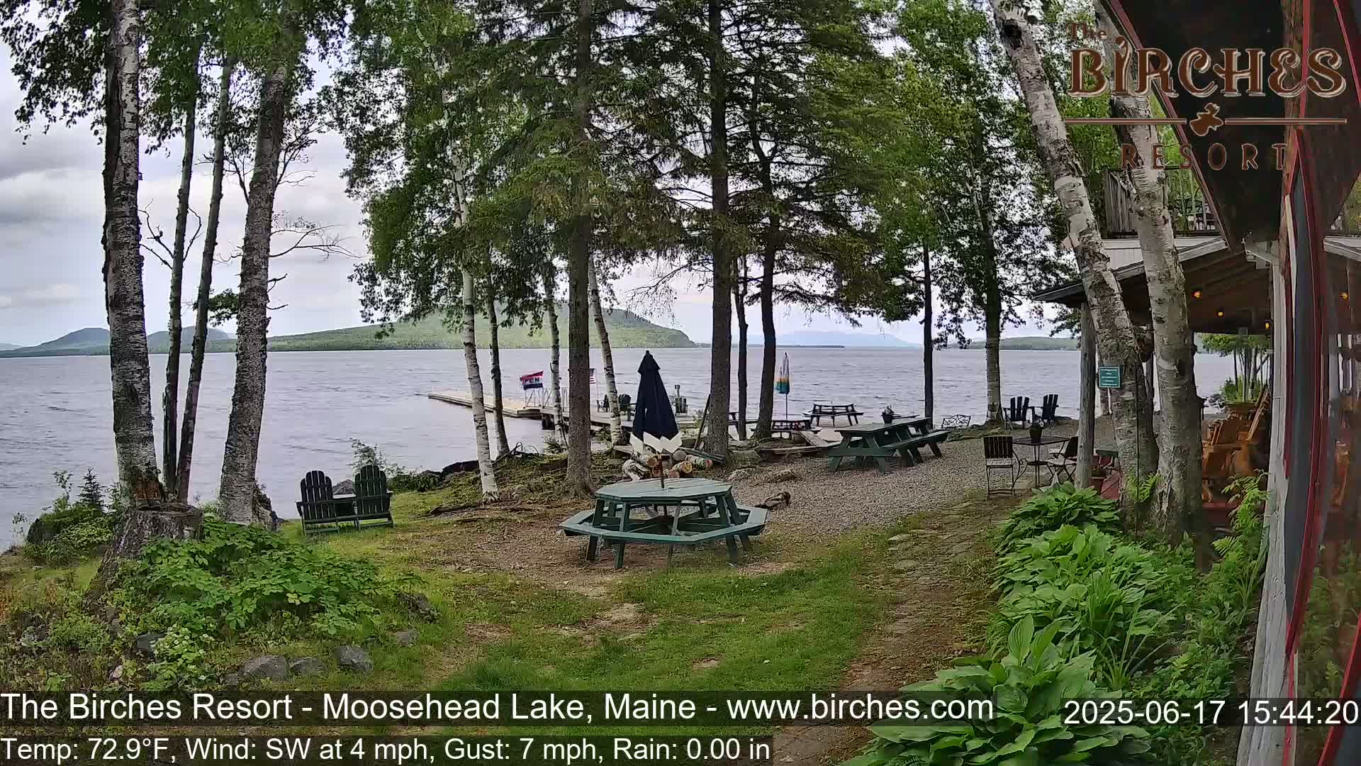 A lakeside patio area with picnic tables and chairs under a partly cloudy sky, nestled among birch trees, overlooks calm water and distant mountains.