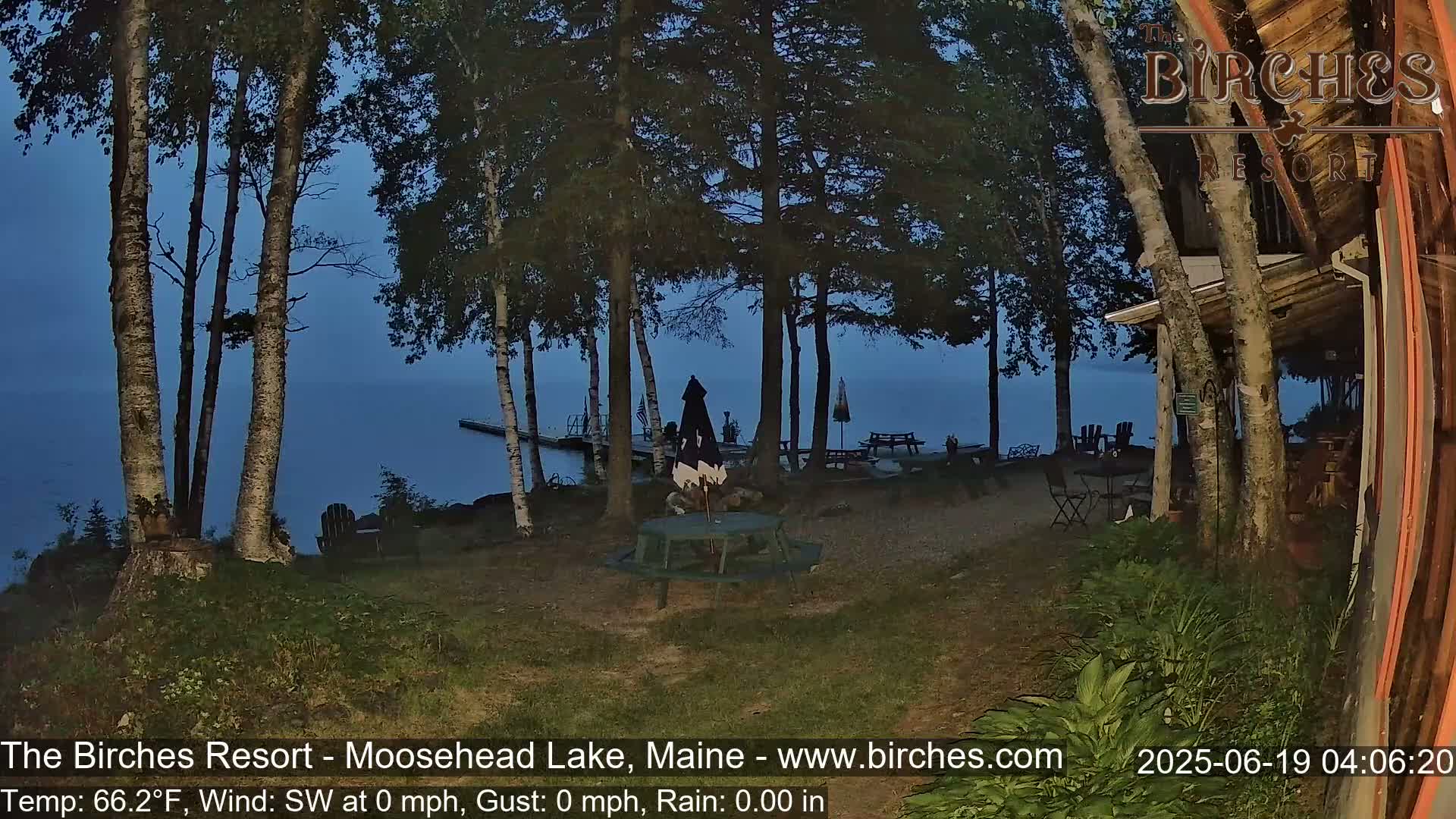 A lakeside resort scene at twilight shows a picnic table and chairs near a calm lake, surrounded by birch trees under a dark blue sky.