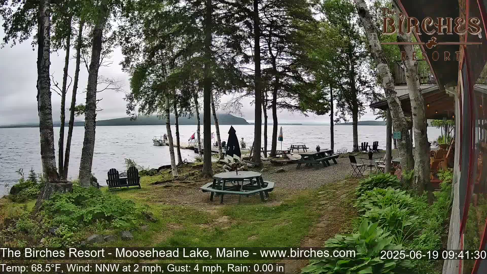 A lakeside patio with picnic tables and chairs is partially shaded by trees on an overcast day with a light wind and rain.