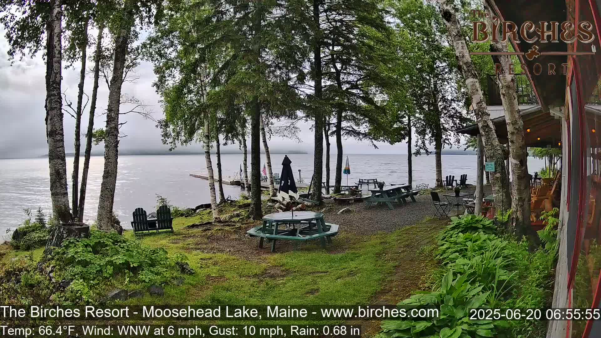 A lakeside patio with picnic tables and chairs under trees in rainy weather, overlooking a calm lake.