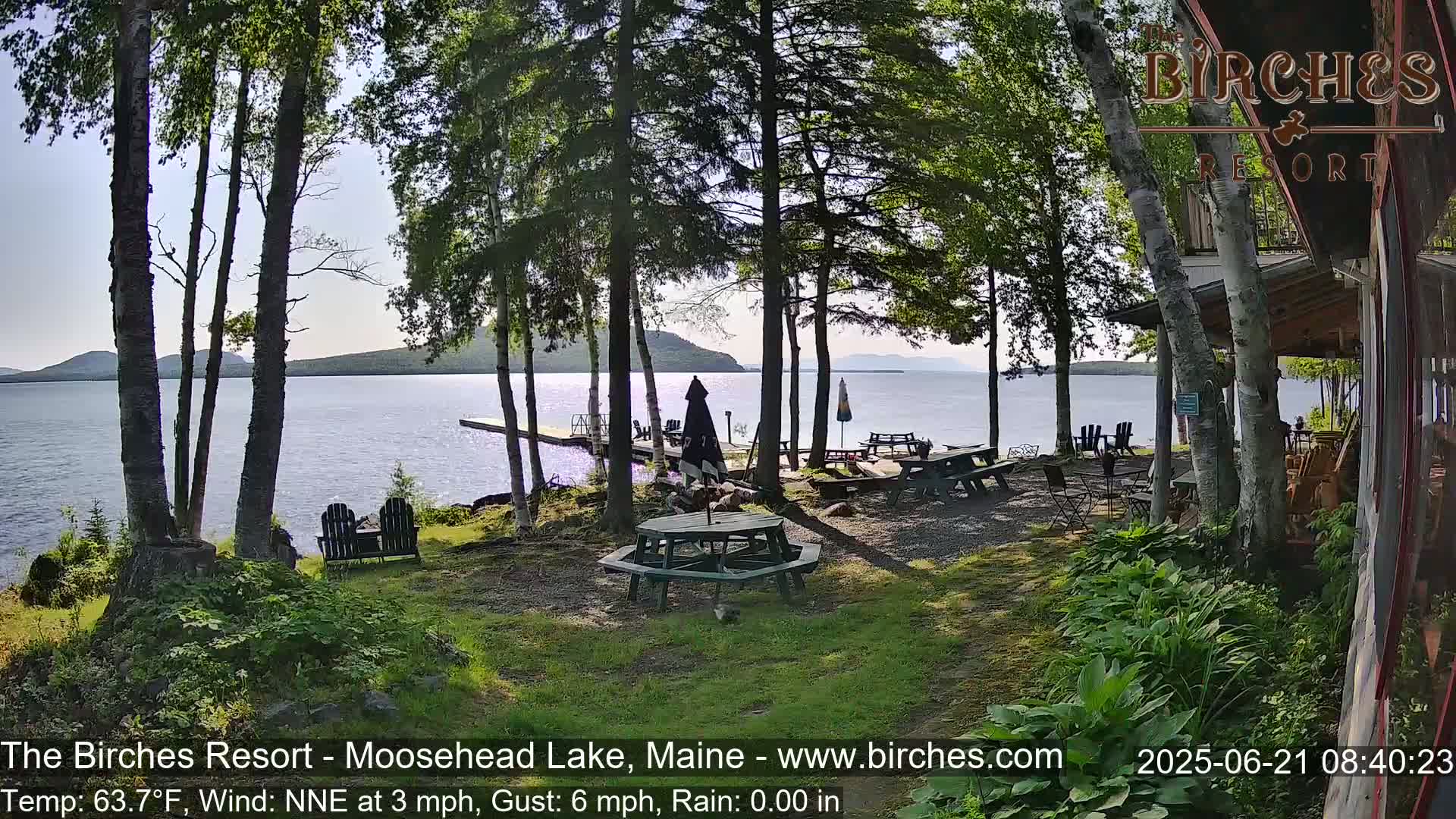 A lakeside patio with picnic tables and chairs sits under a partly sunny sky, surrounded by trees and overlooking calm water.