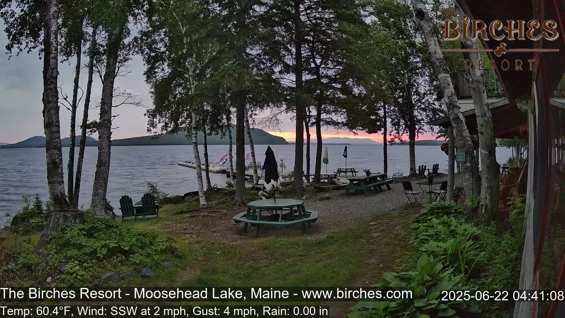 A lakeside patio with picnic tables and chairs sits under a partly cloudy sky at sunset, with light rain falling.