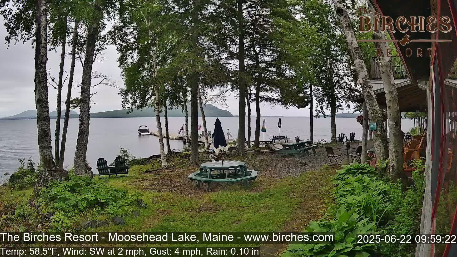 A lakeside resort patio with picnic tables and chairs under a cloudy sky, overlooking a calm lake with a boat docked nearby.