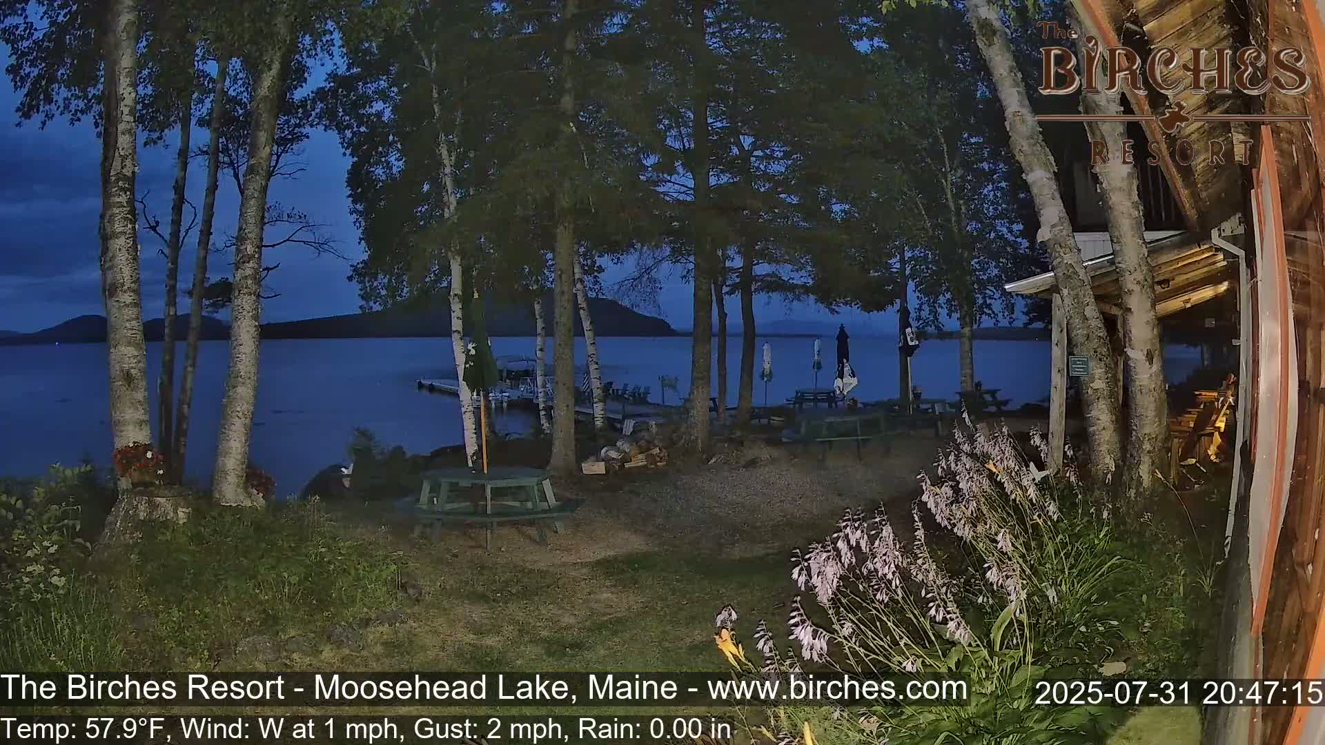 A tranquil lakeside scene at twilight, showing a calm lake with mountains in the distance, viewed from a lakeside property with picnic tables, and a few trees and plants in the foreground under partly cloudy skies.