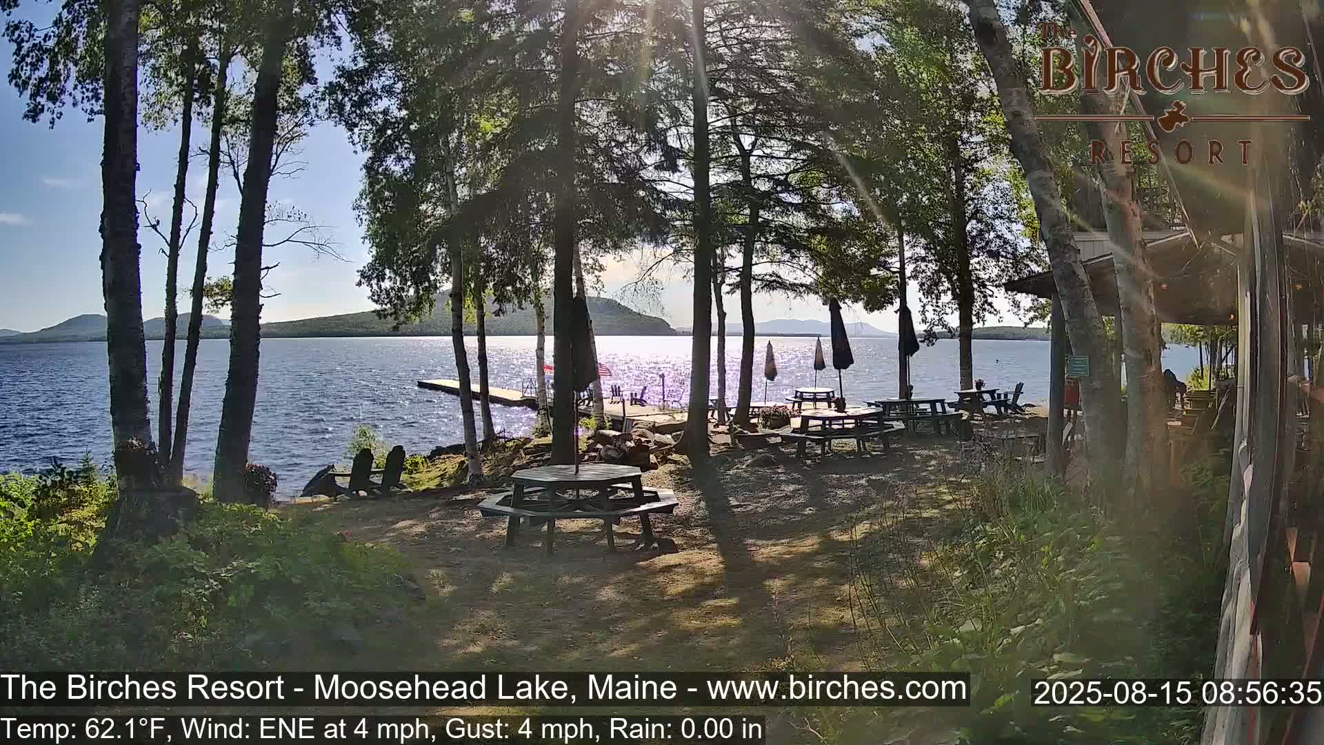 A tranquil lakeside scene at twilight, showing a calm lake with mountains in the distance, viewed from a lakeside property with picnic tables, and a few trees and plants in the foreground under partly cloudy skies.