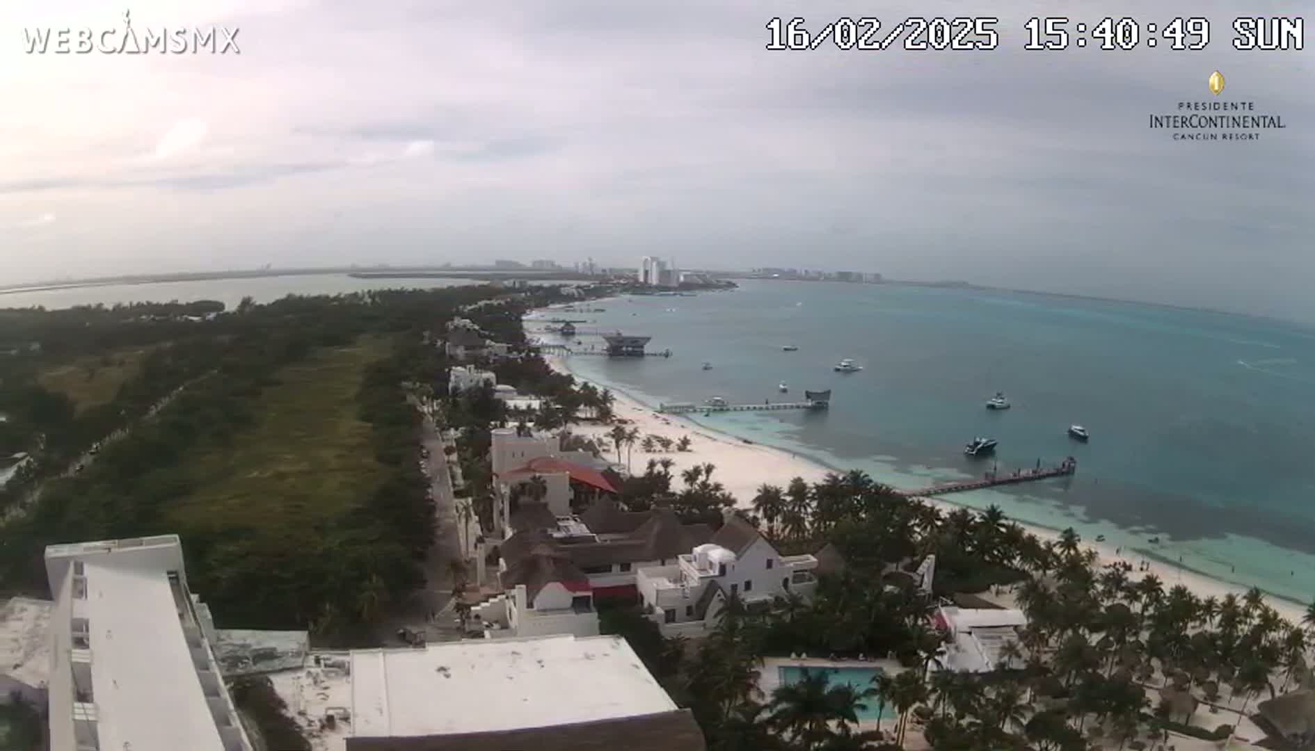 An elevated view shows a partly cloudy day overlooking a white sand beach, turquoise ocean with several boats, and buildings along the coastline, with lush green vegetation inland.