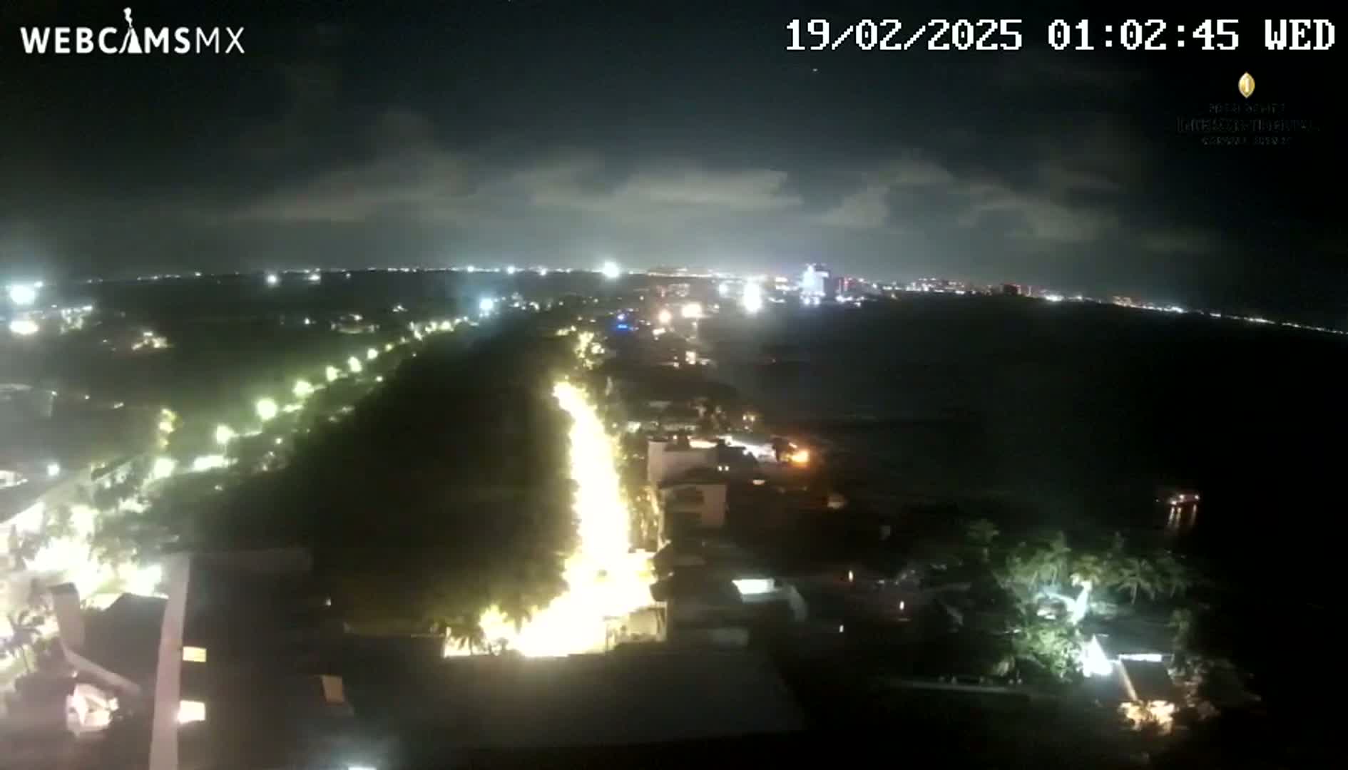 A nighttime, high-angle view shows a brightly lit road winding through a city toward a dark body of water under a partly cloudy sky.