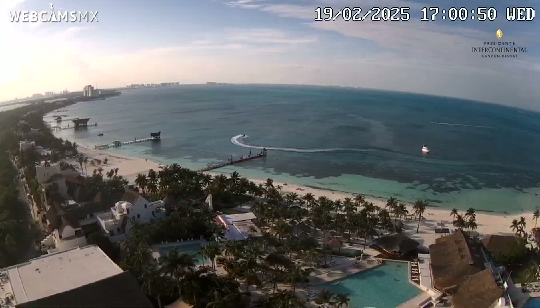 An aerial view shows a tropical beach with turquoise water, palm trees, resort buildings, and a jet ski leaving a wake in the ocean under partly cloudy skies.