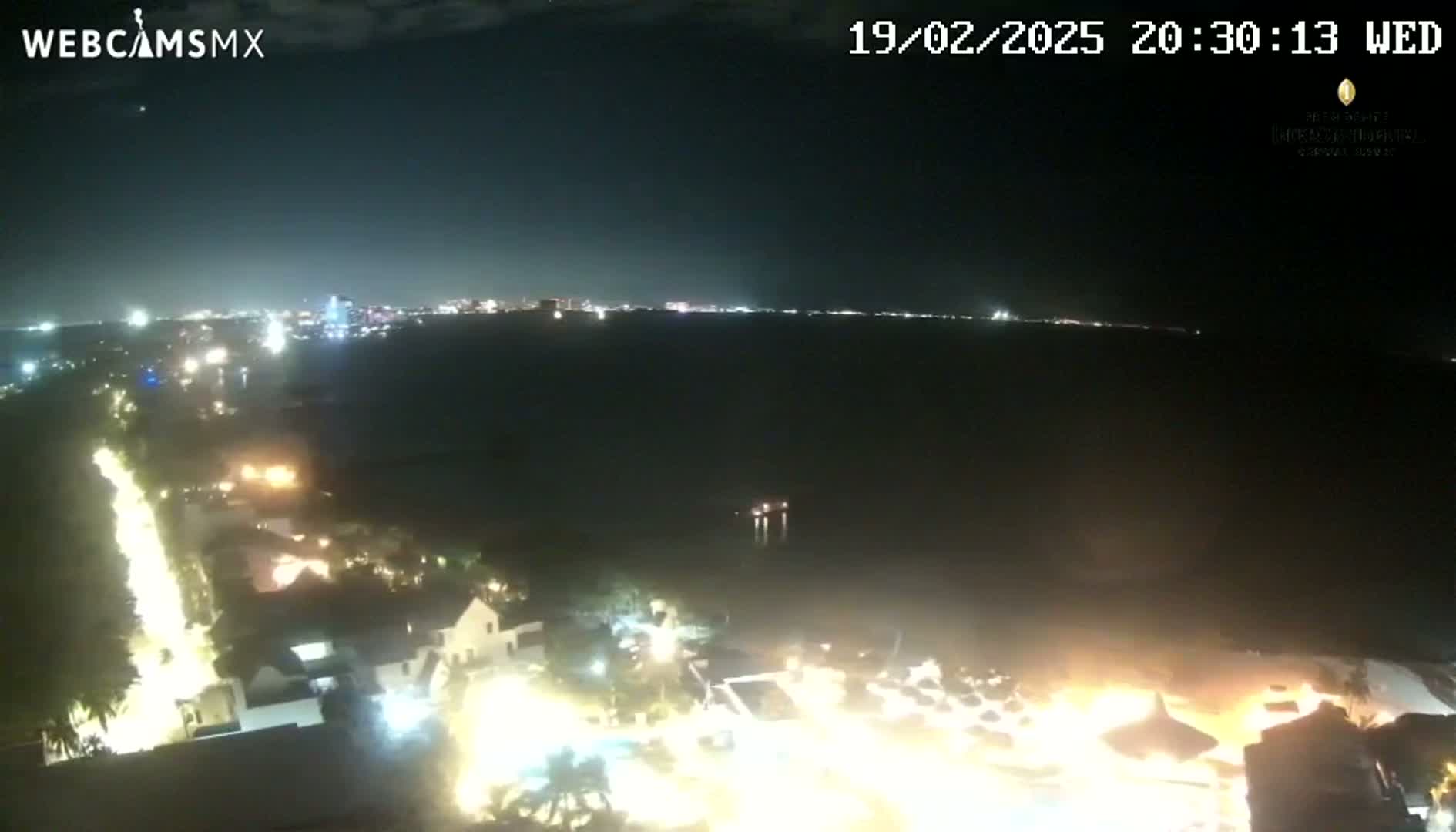 A nighttime aerial view shows a coastal town with brightly lit streets and buildings next to a dark body of water, with a city skyline visible in the distance under a clear sky.
