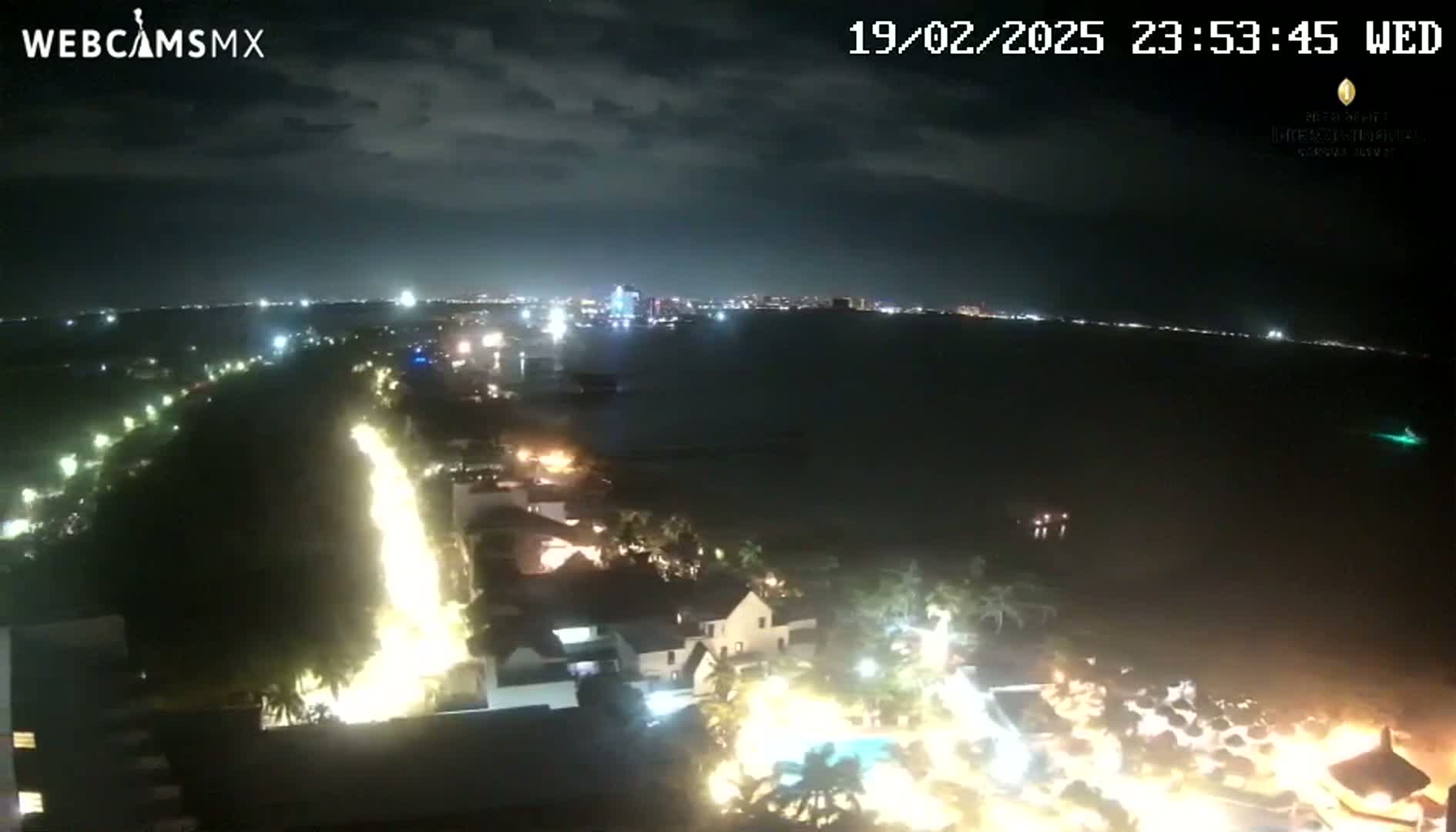 A nighttime, aerial view of a coastal city with brightly lit buildings and roads near a dark body of water under a partly cloudy sky.
