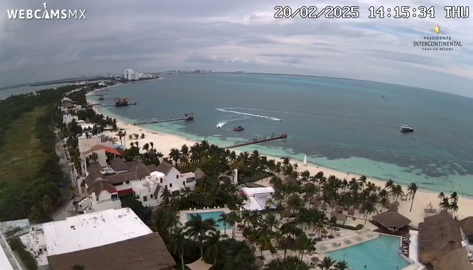 An aerial view shows a tropical resort with swimming pools and palm trees lining a white-sand beach next to a calm, turquoise ocean under a cloudy sky.